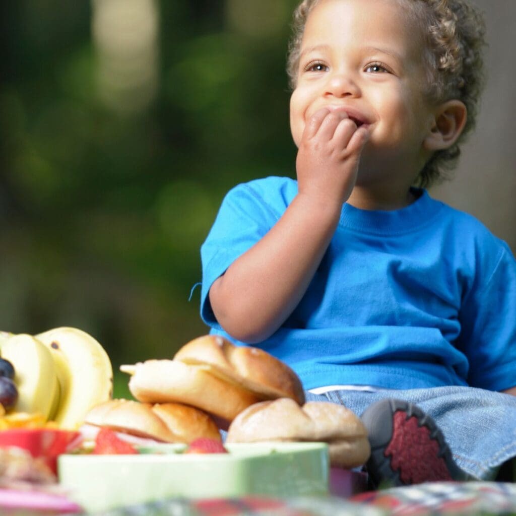 A toddler in a blue tshirt and denim shorts is snacking on something at a picnic. In the foreground are buns, bananas, grapes, and strawberries
