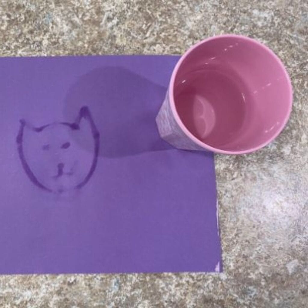 A purple piece of construction paper sitting on the counter with a pink water cup sitting to its side. A paintbrush has been dipped into water and brushed onto the paper to make a cat face.
