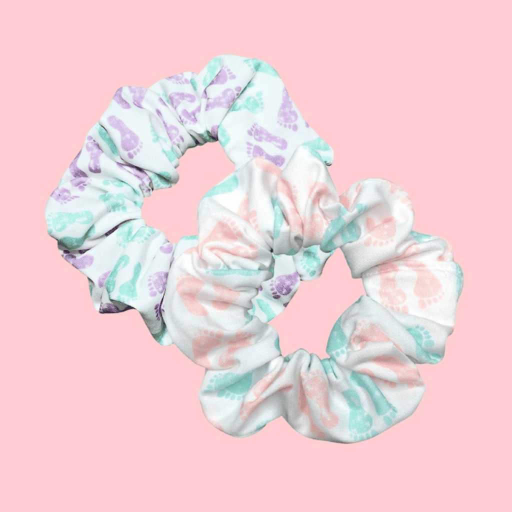 The image shows a fabric hair scrunchie against a soft pink background. The scrunchie has a pastel-colored print with various shades of purple, green, and pink, featuring playful outlines of baby feet prints. Its fabric appears to be soft and possibly of a cotton blend, giving it a cozy, comfortable look suitable for gentle hair styling. The overall aesthetic is cute and would appeal to those who enjoy whimsical or baby-themed accessories.