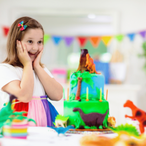 The image shows a joyful young girl at a birthday party, with her hands on her cheeks in a gesture of excitement. She is looking at a dinosaur-themed birthday cake decorated with green icing and dinosaur figurines. The cake is vibrant, featuring layers of bright blue and green, with dinosaur models on top and around the sides. In the background, a colorful string of pennants hangs, adding to the festive atmosphere of the event. The focus is on the girl's delighted expression and the elaborately decorated cake in front of her, illustrating a moment of celebration and happiness at a child's themed party.