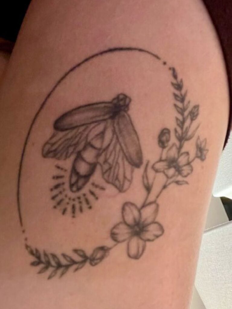 Tattoo on the upper arm of a woman. There is an oval framing a firefly. Half of the oval is made by a simple line with two small dots at each end. The other half of the oval is made with forget-me-nots with leaves trailing away. 