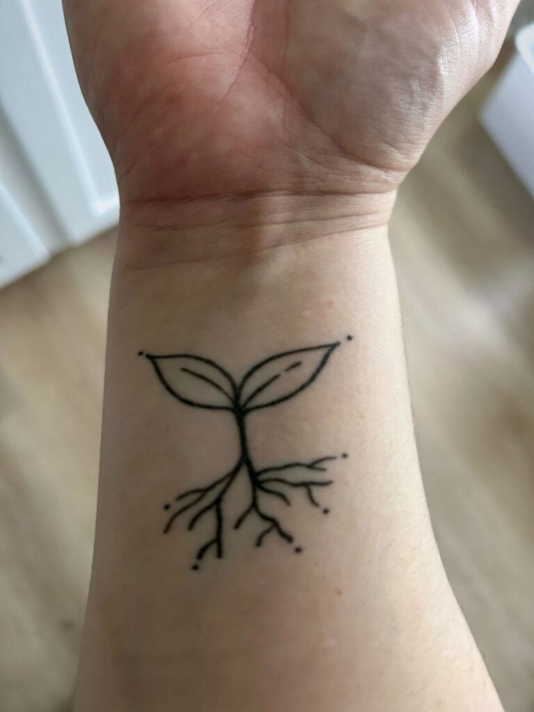 A woman's forearm with a plant sprout tattooed on it. The sprout has two leaves and a small root system