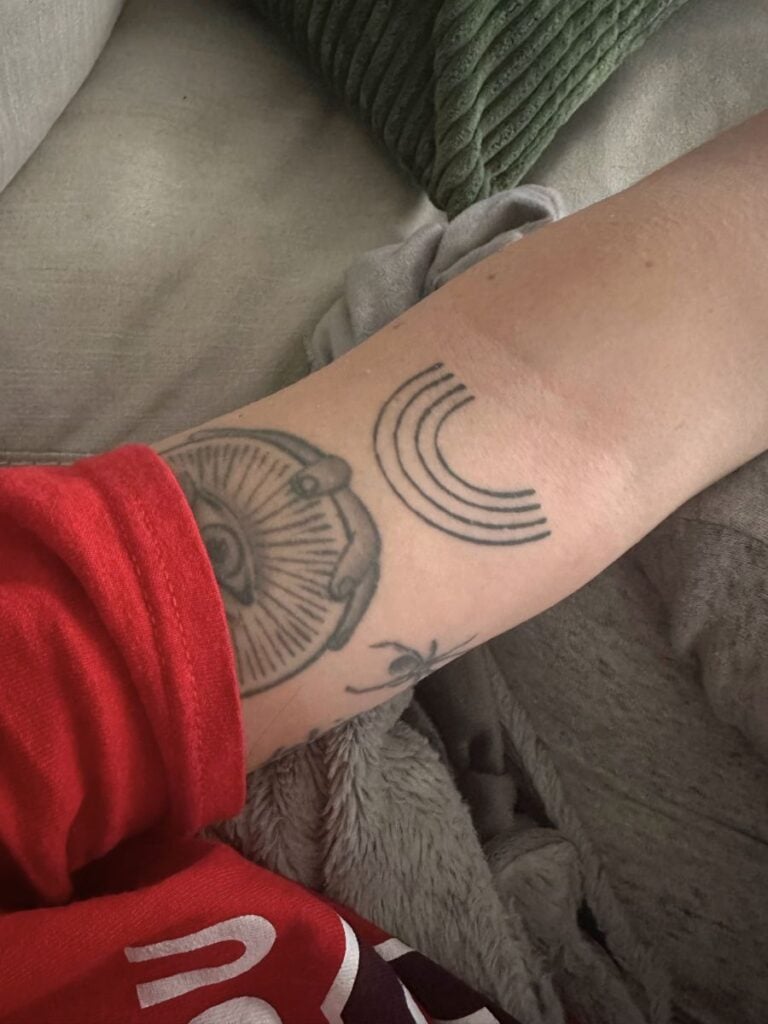 Picture of a mother's arm. Just above the insider of her elbow is a line art tattoo of a black and white rainbow 