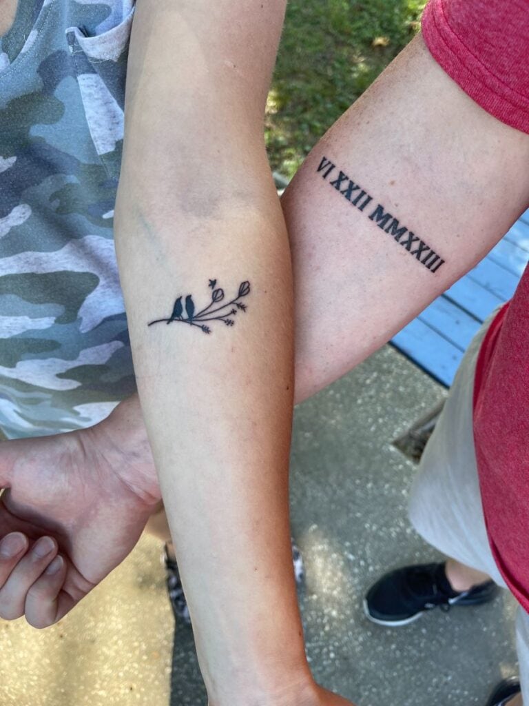 Husband and wife showing their forearms with coordinating tattoos. The wife has a tattoo of two birds sitting on a flowering branch, watching a tiny bird fly away. The husband's tattoo depicts the Roman numerals VI XXII MMXXIII for the date of his wife's miscarriage.