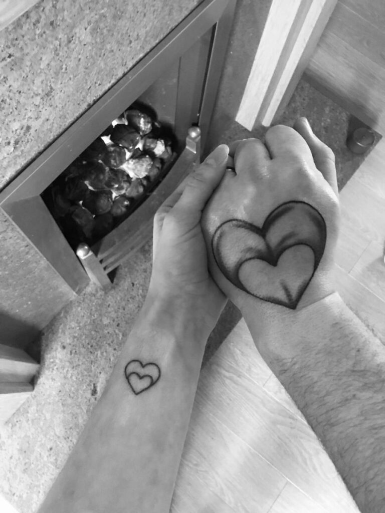 Picture of a woman holding her husband's hand. They each have a tattoo of a heart inside a larger heart. His is on his hand and hers is on her inner wrist. 