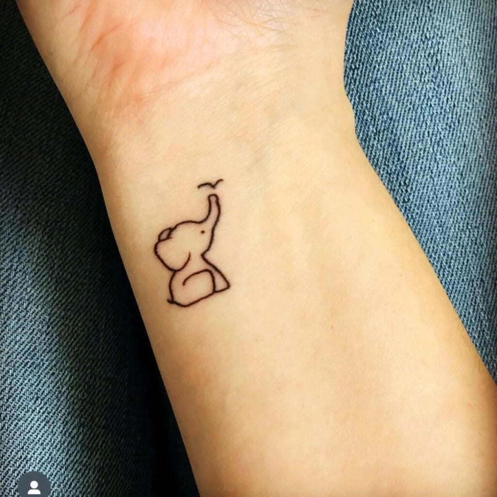 Inner wrist of a woman with a small baby elephant tattoo. The elephant is sitting with her trunk up, and a tiny bird is landing on her trunk