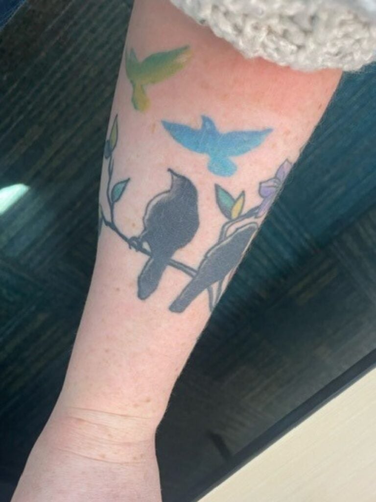 Picture of a woman's forearm with a tattoo. The tattoo shows a branch with two black birds' silhouettes facing away and watching two birds (vivid green and bright blue) fly away. Wrapped around past where can be seen is a third bird sitting on the branch.