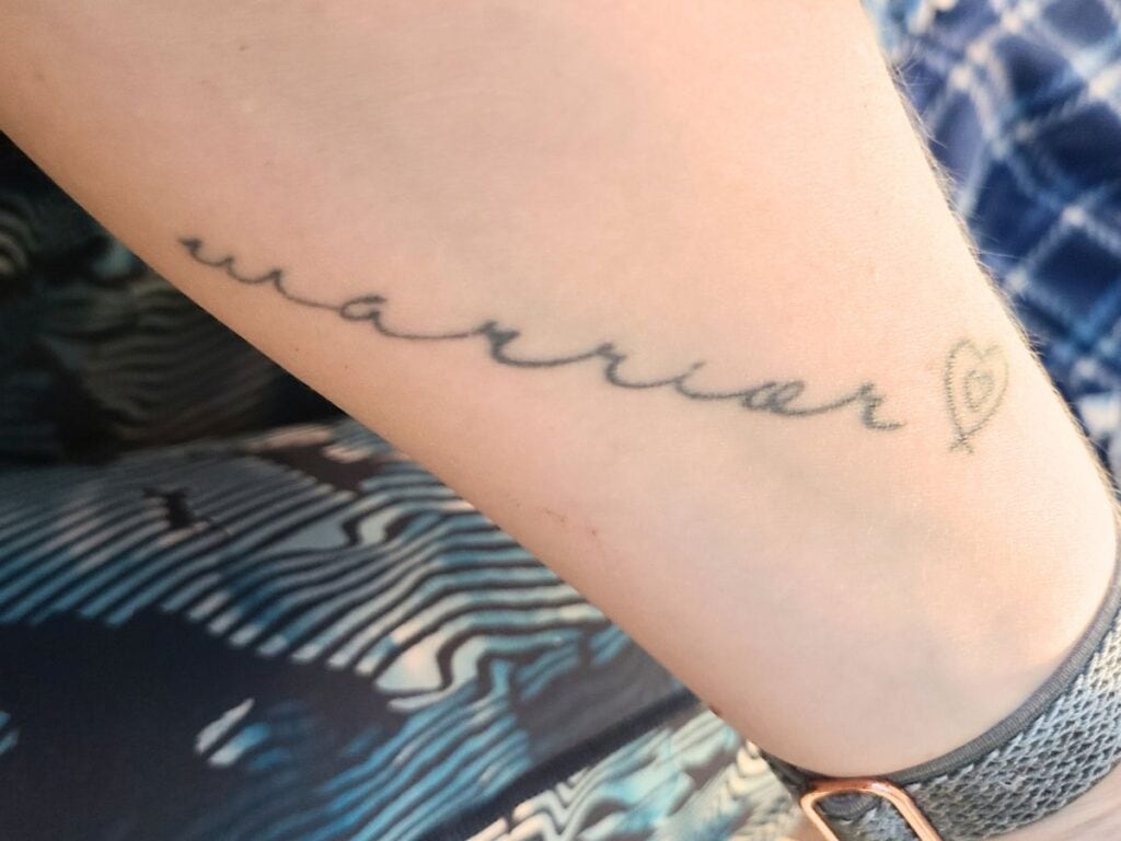 Woman's forearm with the word "warrior" tattooed in a wide script. Beside it is line art of one small heart inside another.