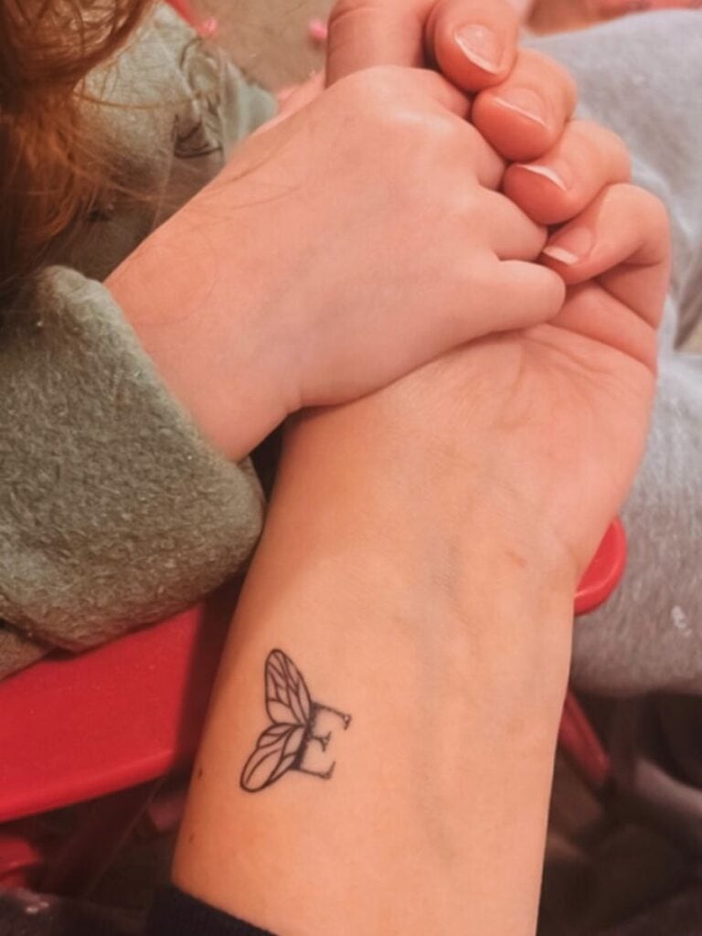A picture of a mother holding her young daughter's hand. On the inside of the mother's wrist is a tattoo: An "E" with small butterfly wing attached to the left side of its post