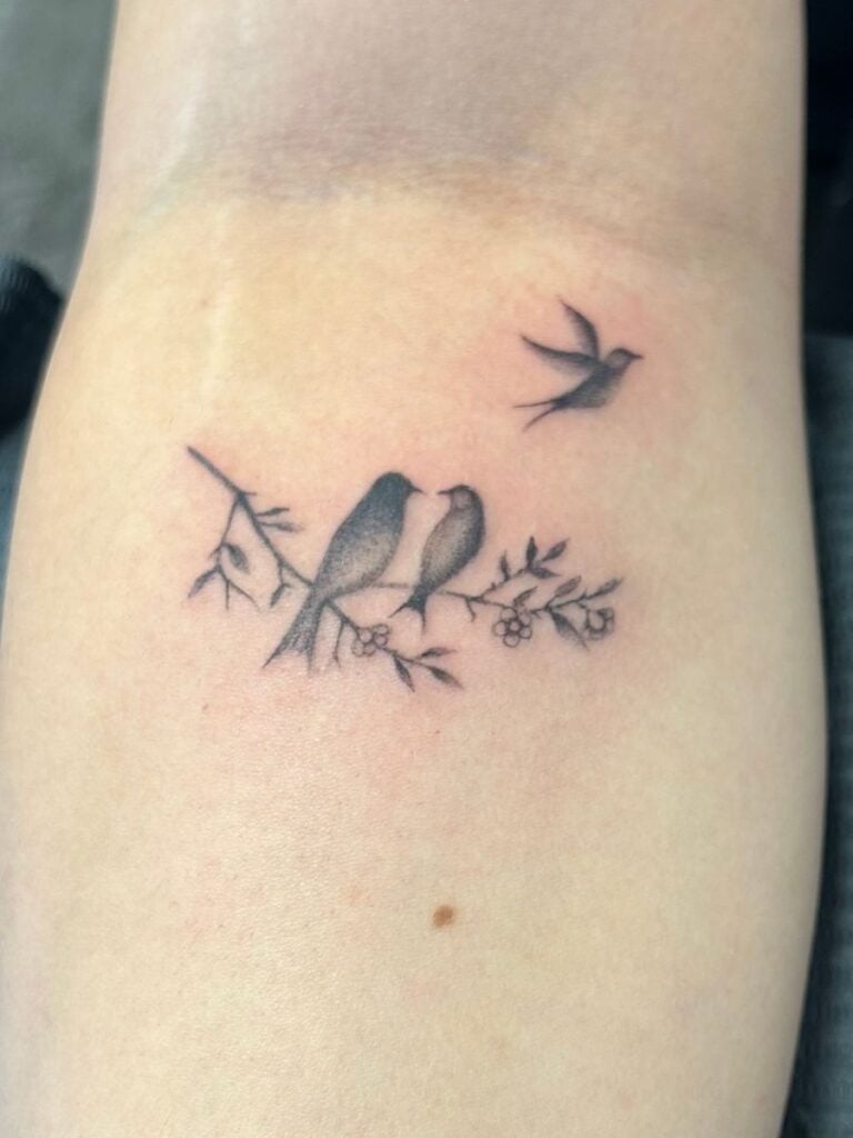 A tattoo on a mother's forearm of two birds sitting on a flowering branch looking at one another while a third bird flies away.