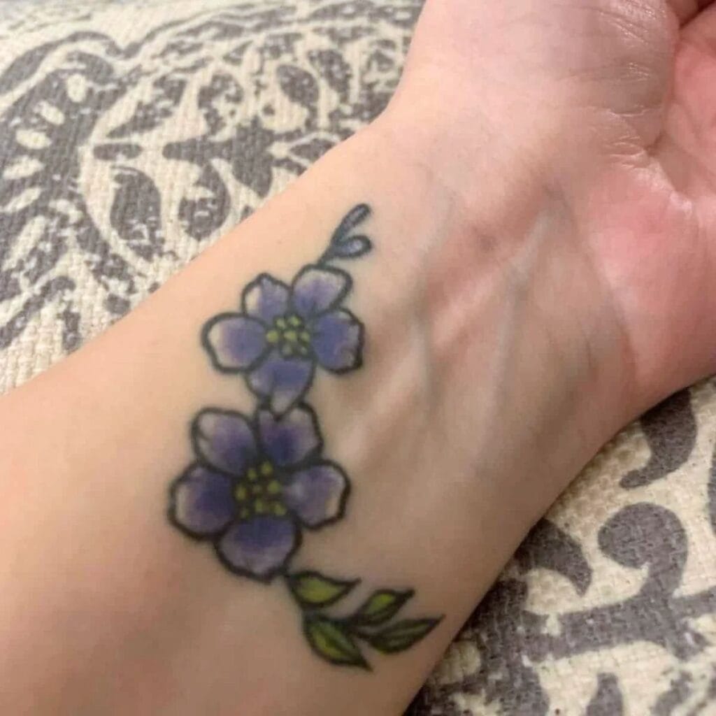 Tattoo at the inside of a mother's wrist showing forget-me-nots. The blossoms are purple with yellow centers, and there are a few green leaves to the side.