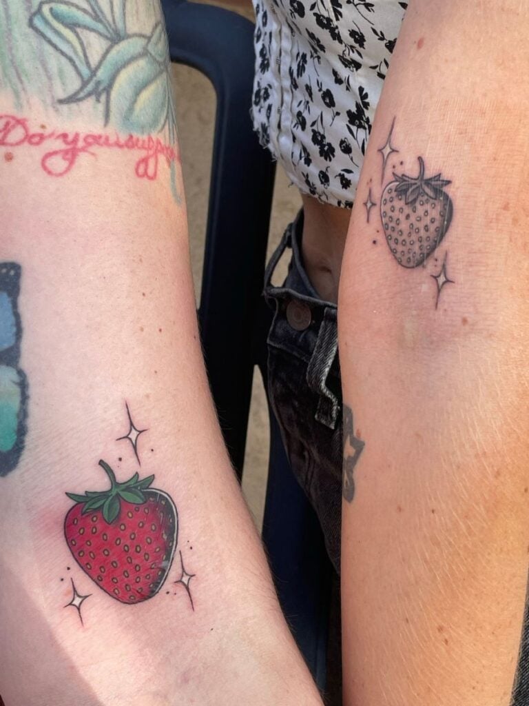 Picture of two romantic partners' arms. Each has a strawberry tattooed on their upper arm, although one is colored in while the other is black and white. 