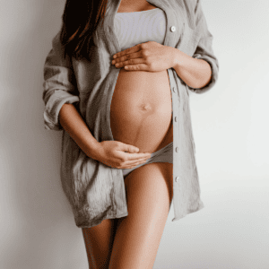 An expectant mother in her third trimester poses for a maternity photo. She stands against a neutral background, wearing a lilac tank top and grey maternity underwear. Her belly is lovingly cradled by her hands, emphasizing her baby bump. An open, long-sleeved grey shirt adds a casual layer to her outfit, suggesting a comfortable, intimate setting.
