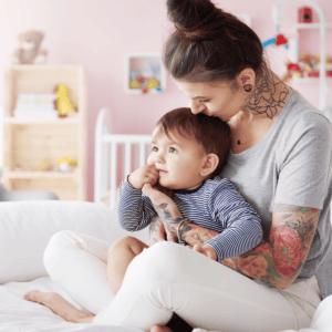 An affectionate moment between a mother and her toddler in a softly lit nursery room. The mother, sporting a casual bun and a gray t-shirt, has several vibrant tattoos along her arms and neck, featuring a mix of floral and abstract designs. She gently embraces her young child, who is gazing off into the distance with a thoughtful expression. The child, dressed in a navy and white striped top, sits comfortably in the mother's lap. The background reveals a well-kept nursery, with toys neatly arranged on shelves and a cozy ambiance.
