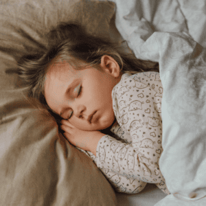 A peaceful toddler sleeps soundly, nestled in soft bedding. The child, likely between two to three years old, is wearing a long-sleeved pajama set adorned with a cute, light-colored animal print. The youngster's cheek rests gently against a folded arm, while tousled hair adds a touch of innocence to the serene scene. A taupe pillow and light gray comforter offer a warm and comforting backdrop, evoking a sense of tranquility and restfulness.